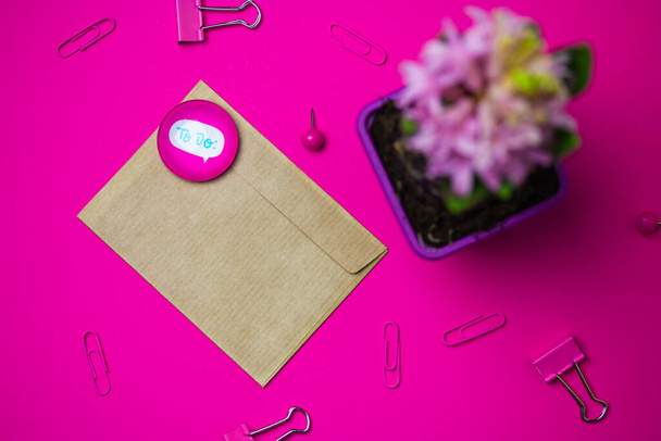 Bruine envelop op roze achtergrond van boven. Papierwaren - roze papieren bindmiddelen, paperclips. Potted lente hyacint bloem in bloei op tafel Vrouw bureau met een letter, magneet stapel met tekst te doen - Foto, afbeelding