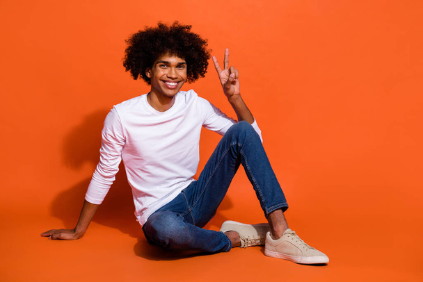 Photo of cute attractive guy wear white shirt showing v-sign sitting floor isolated orange color background - Фото, изображение