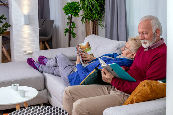 Sevgili yaşlı çift dinleniyor ve evdeki kanepede kitap okuyor. Emeklilik günlerinde boş zaman. - Fotoğraf, Görsel