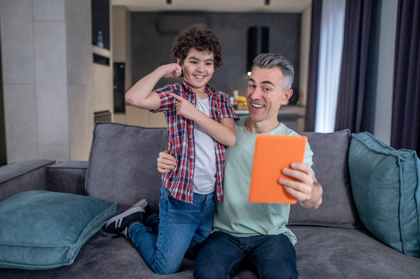 Boy showing arm muscles and smiling dad with tablet - Φωτογραφία, εικόνα