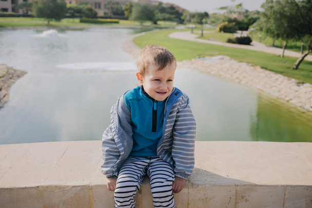 Άτακτο παιχνιδιάρικο παιδί του νηπιαγωγείου, κάθεται στη γέφυρα κοντά στη λίμνη. - Φωτογραφία, εικόνα