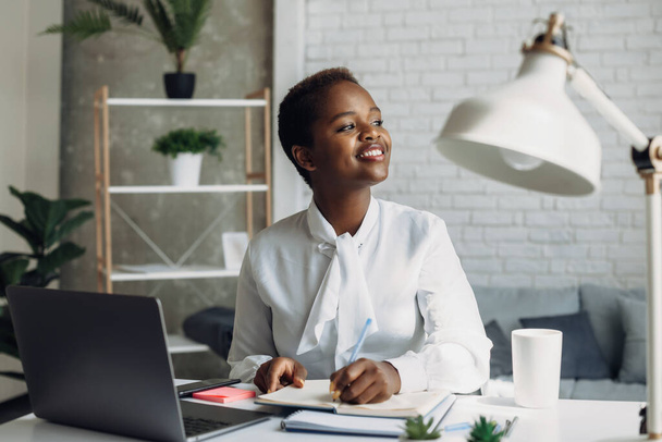 Hymyilevä musta toimisto nainen istuu läppärissä tekemässä muistiinpanoja töissä. Internet-teknologia. Afroamerikkalainen tyttö. - Valokuva, kuva