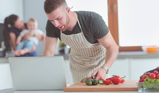 Young man cutting vegetables and woman standing with laptop in the kitchen - Fotó, kép