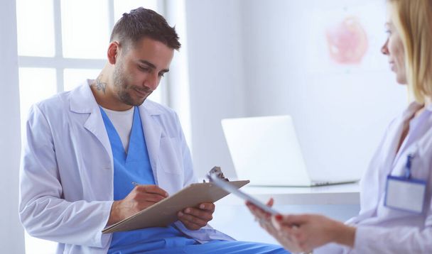 Handsome doctor is talking with young female patient and making notes while sitting in his office - Photo, image