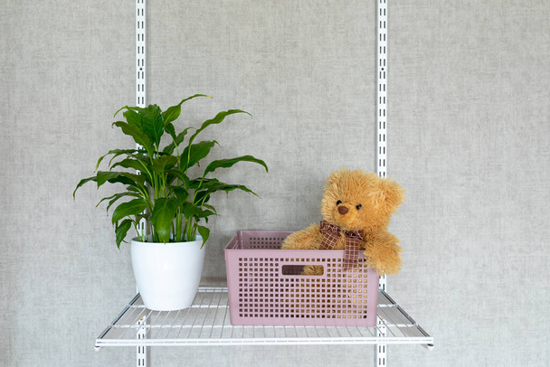 Un jouet d'ours en peluche dans un panier en plastique et un spathiphyllum de plantes d'intérieur sur une étagère suspendue en filet. Systèmes de rangement modernes dans la chambre des enfants. - Photo, image