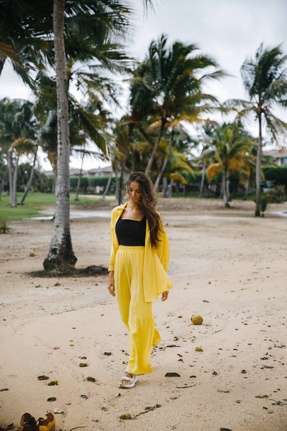 a beautiful skinny brunette girl in a yellow suit poses on vacation near tropical palm trees at a golf club in the Dominican Republic - Zdjęcie, obraz
