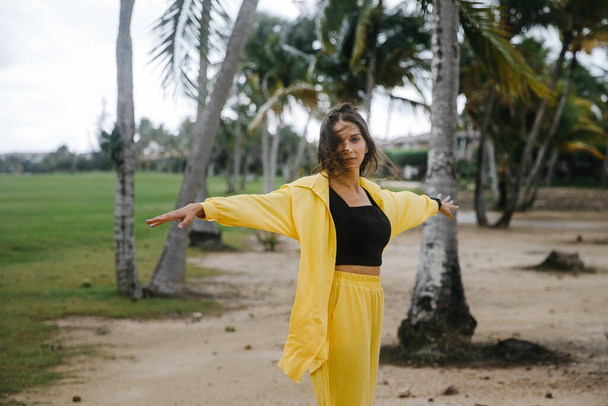 a beautiful skinny brunette girl in a yellow suit poses on vacation near tropical palm trees at a golf club in the Dominican Republic - Fotografie, Obrázek