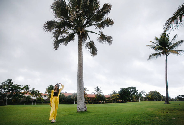 a beautiful skinny brunette girl in a yellow suit poses on vacation near tropical palm trees at a golf club in the Dominican Republic - Fotoğraf, Görsel