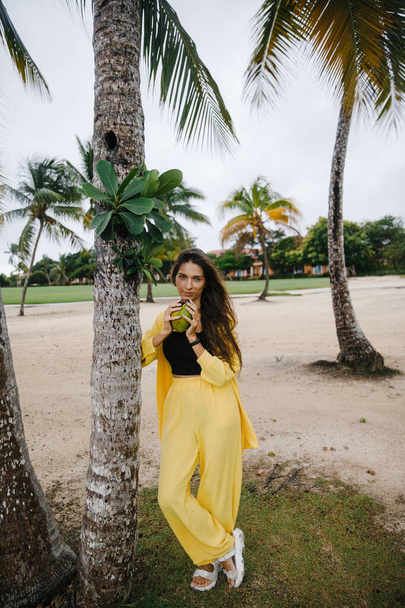a beautiful skinny brunette girl in a yellow suit poses with a coconut on vacation near tropical palm trees at a golf club in the Dominican Republic - Foto, Bild