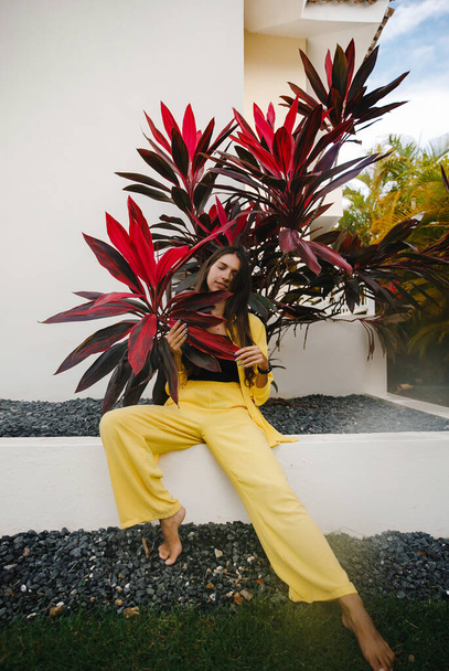 a beautiful skinny brunette girl in a yellow suit and a black top poses on vacation near tropical red leaves at a golf club in the Dominican Republic - Foto, afbeelding