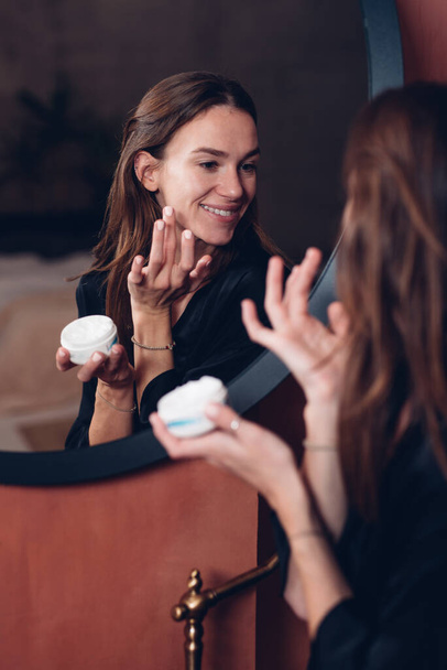Mujer joven delante del espejo poniéndose crema en la cara - Foto, Imagen