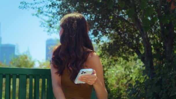 Mulher asiática olhando para trás na paisagem urbana borrada segurando smartphone no parque close-up - Filmagem, Vídeo