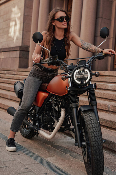 スタイリッシュな女性ポージングオン古典的なダークバイク屋外 - 写真・画像