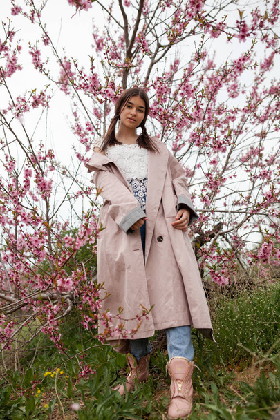 beautiful girl in spring peach garden. Teenager dressed in raincoat, jeans - Φωτογραφία, εικόνα
