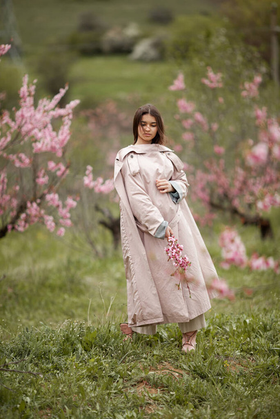 beautiful girl in spring peach garden. Teenager dressed in raincoat, jeans - Photo, image