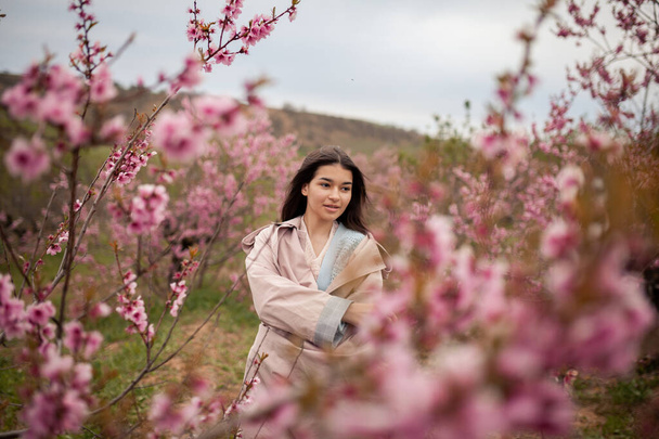 beautiful girl in spring peach garden. Teenager dressed in raincoat, jeans - Foto, Imagen