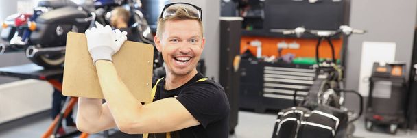 Hombre feliz reparador sujetando portapapeles con documentos cerca de la motocicleta en el taller de reparación de automóviles - Foto, Imagen