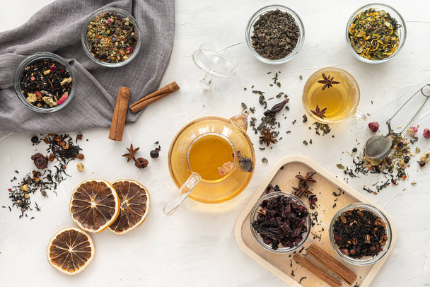Several varieties of herbal and fruit tea in glass bowls and a kettle with a cup of hot tea on a light background. View from above. Healthy eating. Delicious warming drink.  - Zdjęcie, obraz