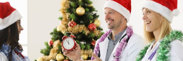 Ärzte in Weihnachtsmützen halten Wecker und Neujahrsgeschenke in der Nähe des Weihnachtsbaums - Foto, Bild