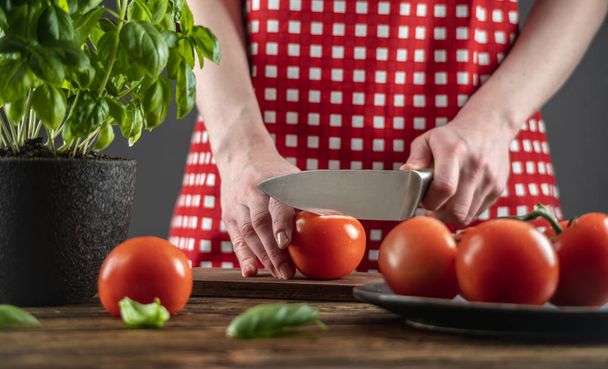Chef in a red apron is cutting tomatoes, basil in a pot is on the table next to it. Process of cooking dish from fresh vegetables, healthy food. - Photo, image