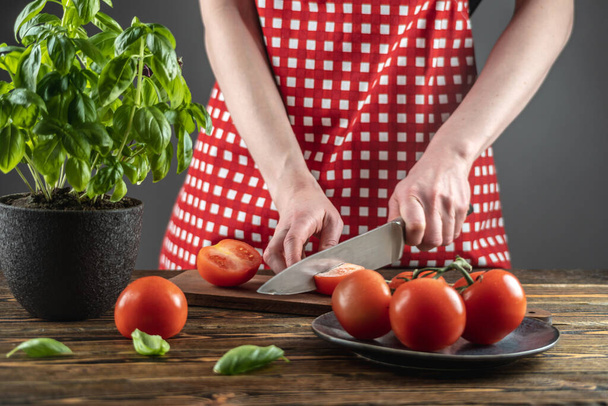 Woman in a red apron is cutting tomatoes, basil in a pot is on the table next to it. Process of cooking dish from fresh vegetables, healthy food. - Photo, image