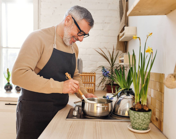 Счастливый пожилой человек в фартуке готовит дома на кухне, стоит рядом с плитой и перемешивает соус в кастрюле - Фото, изображение