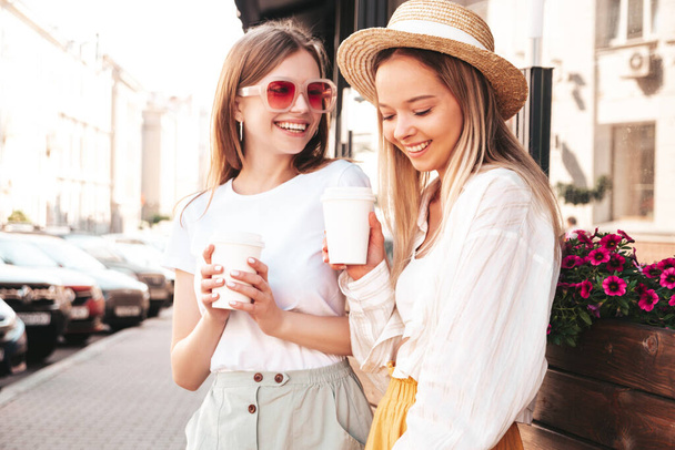 Two young beautiful smiling hipster female in trendy summer clothes.Sexy carefree women posing in the street. Positive pure models having fun at sunset. They drinking coffee or tea in plastic cup - Foto, afbeelding
