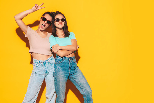 Two young beautiful smiling blond hipster female in trendy summer clothes. Sexy carefree women posing near yellow wall in studio. Positive models having fun. Cheerful and happy. In sunglasses - Photo, image