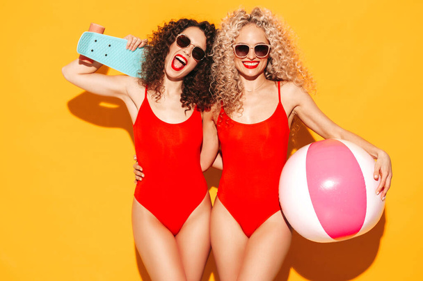 Two beautiful sexy smiling hipster women in red summer swimwear bathing suits.Trendy models with afro curls hairstyle having fun in studio.Hot female isolated on yellow.Holding penny skateboard - Photo, Image