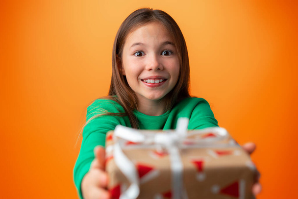 Cute teen girl holding a Christmas gift box against orange background. - Foto, afbeelding