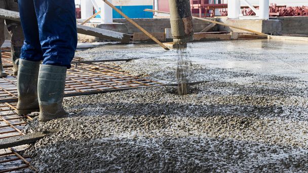 Строители заливают бетонный пол в промышленный цех. Ноги в сапогах в бетоне. Подача бетона для заливки пола. Монолитный бетонный завод - Фото, изображение