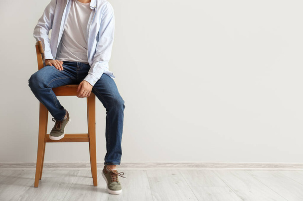 Fashionable young man in stylish jeans sitting on chair near light wall - Foto, Imagem