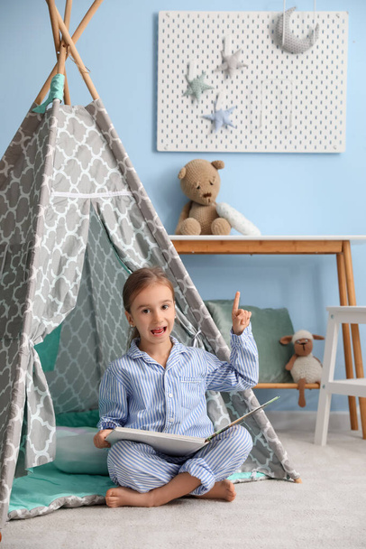Linda niña en pijama con libro apuntando al tablero de clavijas colgantes en la pared azul - Foto, Imagen