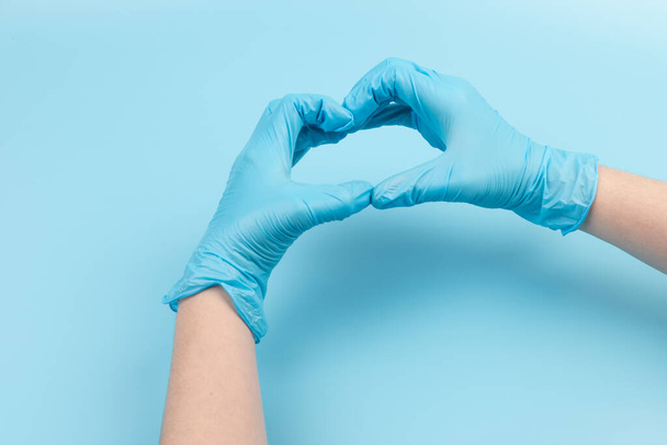 Persona en guantes de látex mostrando el gesto del corazón contra el fondo azul claro, primer plano en las manos - Foto, Imagen