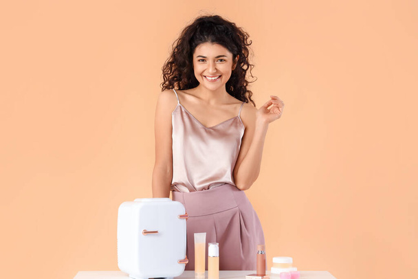 Красива усміхнена жінка стоїть біля столу з холодильником і косметичними продуктами на кольоровому фоні
 - Фото, зображення