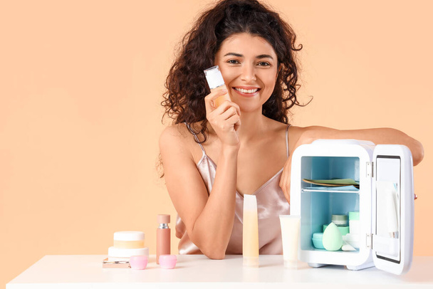 Jonge vrouw in de buurt van tafel met geopende koelkast en cosmetische producten op kleur achtergrond - Foto, afbeelding
