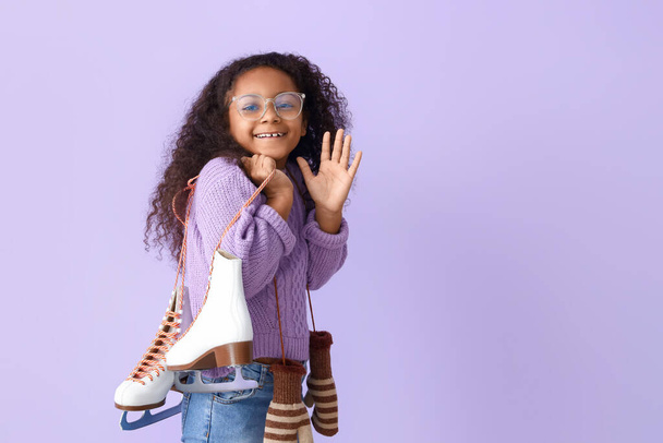 Little African-American girl in eyeglasses with ice skates waving hand on lilac background - Foto, immagini