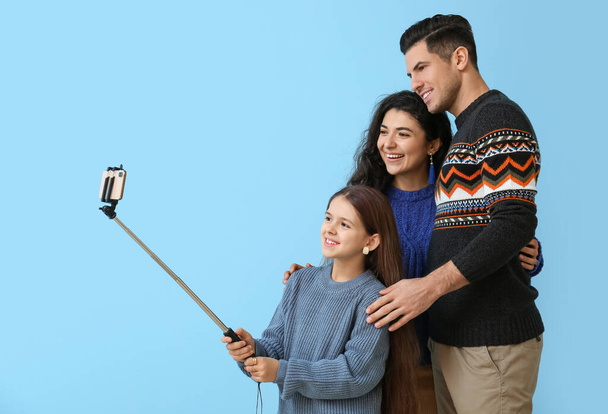 Little girl and her parents in warm sweaters taking selfie on blue background - Foto, imagen