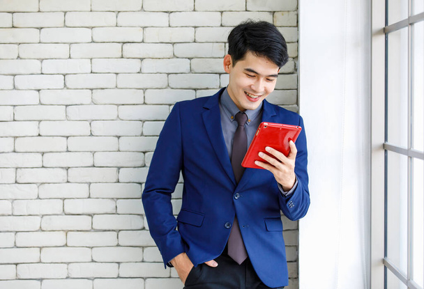 Retrato estudio disparo de asiático guapo profesional exitoso empresario masculino en traje azul formal corbata de pie sosteniendo teléfono inteligente sonriendo sobre fondo de pared de ladrillo blanco. - Foto, Imagen