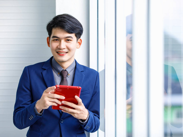 Millennial asiático guapo inteligente seguro de éxito profesional gerente masculino empresario en traje azul formal sonriendo sosteniendo teléfono inteligente de pie cerca de la ventana de la oficina de rascacielos - Foto, Imagen