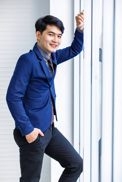 Миллениальный азиатский красивый умный уверенный в себе профессиональный успех мужчина менеджер бизнесмен предприниматель в формальном синем костюме улыбается стоя у окна офиса небоскреба - Фото, изображение
