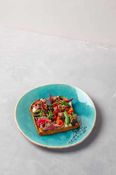 Bruschetta with roast beef, baked paprika and onion on blue ceramic plate. Toast with roast beef in contemporary style. Chef concept bruschetta in modern ceramic dishware. Handmade dinner plate - Photo, Image