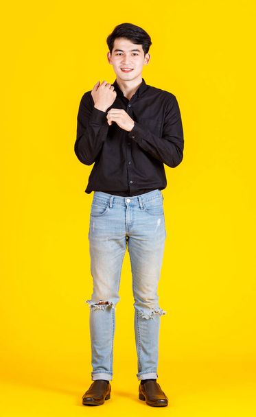 Portrait studio shot of millennial Asian professional successful male businessman in casual black long sleeve shirt outfit standing holding hand in tearing denim jeans pocket on yellow background. - Photo, Image