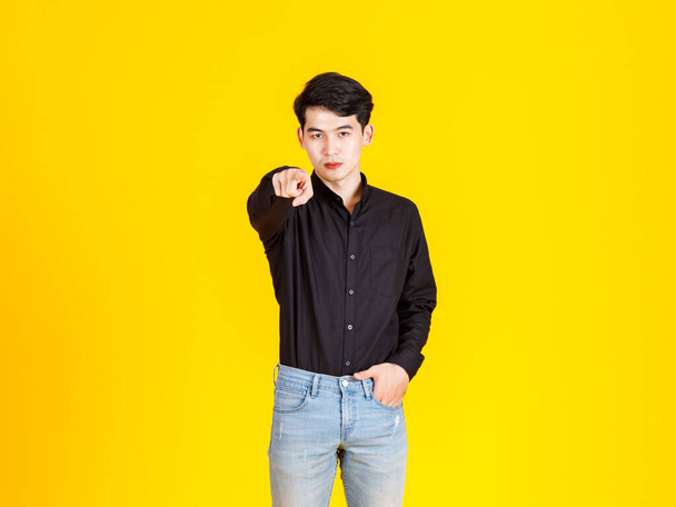 Portré stúdió lövés millenniumi ázsiai profi sikeres férfi üzletember alkalmi fekete ing álló kéz farmerzsebben mutatja hüvelykujj fel mosolygós pillantást kamera sárga háttér. - Fotó, kép