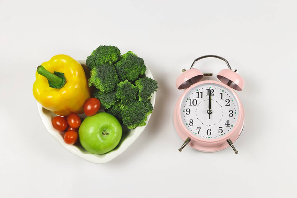 Bovenaanzicht of platte lay van groenten capsicum, broccoli en tomaat in hartvorm plaat op witte achtergrond met roze vintage wekker, gezonde levensstijl en intermitten vasten concept. - Foto, afbeelding