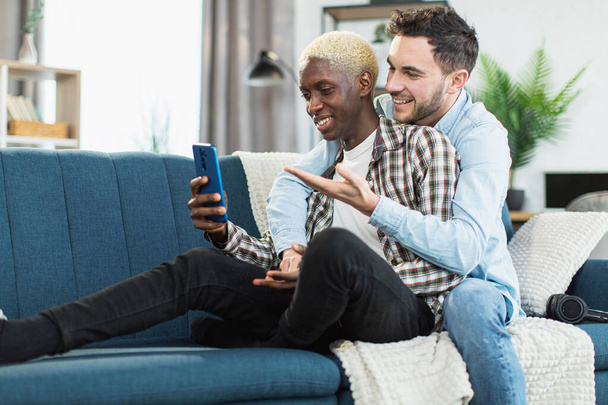 Diverse gays sitting in embrace on couch and using mobile - Foto, Bild