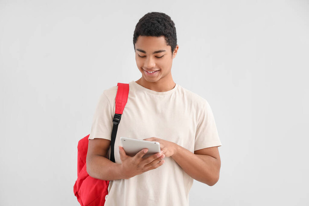 Male African-American student with tablet computer and backpack on light background - Fotó, kép