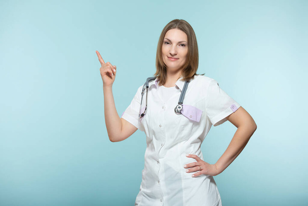 A beautiful female doctor with a stethoscope points to the place where there will be important information, on a blue background. Copy paste. healthcare concept. - 写真・画像