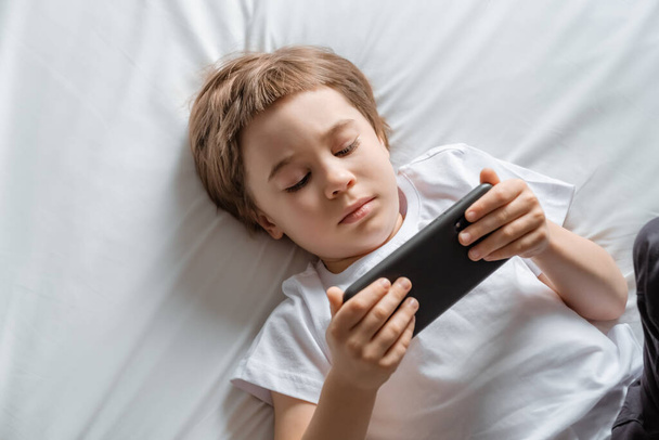 Little baby boy lays on his bed and plays with a smartphone. Close-up portrait. Holds in hands black cell phone. Concept of online education. White room and clothes. Telephone user. Save child vision - Fotoğraf, Görsel