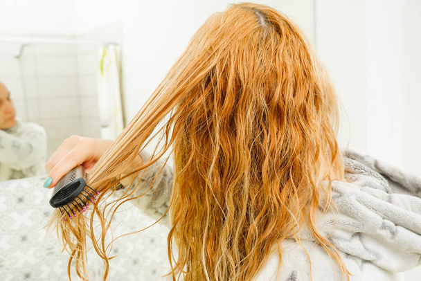 Μια ξανθιά γυναίκα προσπαθεί να χτενίσει τα μπερδεμένα μαλλιά της με μια χτένα μετά το μπάνιο. Κόκκινο ξανθό χτένισμα βρεγμένα και μπερδεμένα μαλλιά. - Φωτογραφία, εικόνα